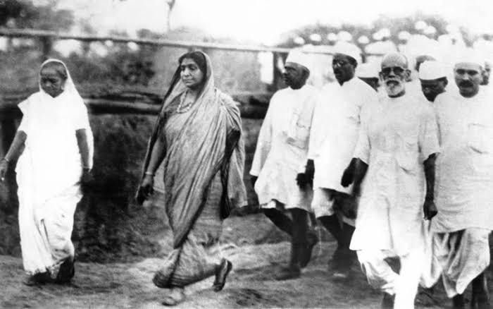 Sarojini Naidu walking in a group of marchers on an open road, with a line of men in white khadi caps behind.