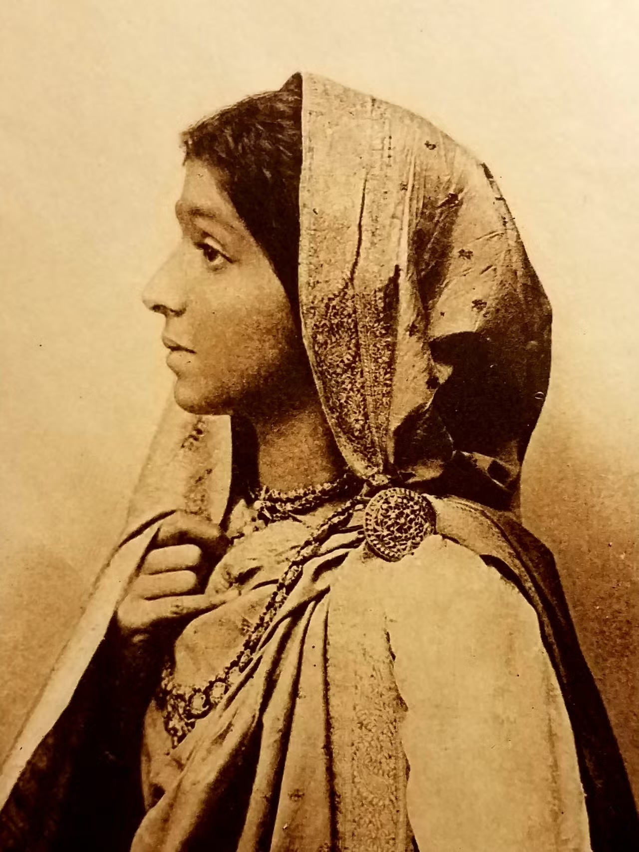 Sepia studio portrait of a young Sarojini Naidu in profile, wearing an embroidered sari drawn over her hair.