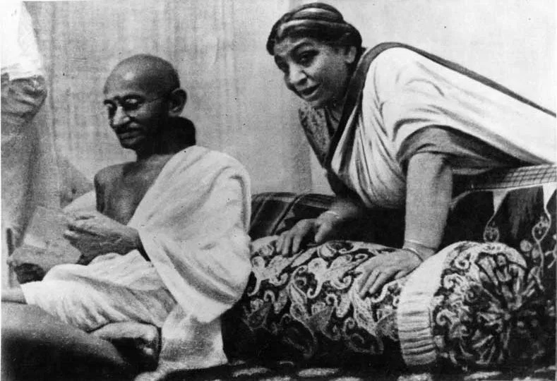 Mahatma Gandhi seated cross-legged on the floor beside Sarojini Naidu, who leans forward on a low seat, mid-conversation.