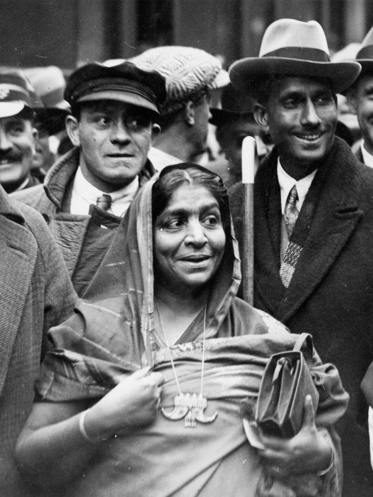Sarojini Naidu at the centre of a dense crowd of men in overcoats and hats on a London street.